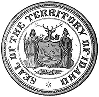 A round seal with the words 'seal of the territory of Idaho' around the outside edge and an image of two figures on either side of a shield painted with a landscape of mountains and rivers. Above the shield is the head of a deer with large antlers.