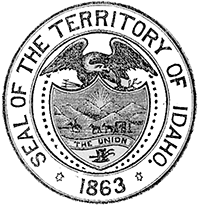 A round seal with the words 'seal of the territory of Idaho 1863' around the outside edge and an image of a bald eagle on top of a shield painted with a landscape people on horses and a carriage in front of mountains and a sunrise with a banner that says 'the union'.